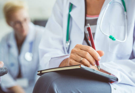 A medical professional sitting with a notebook on her lap and a stethoscope around her neck.