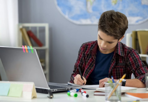 A young student taking notes in a notebook from information gained on his laptop.