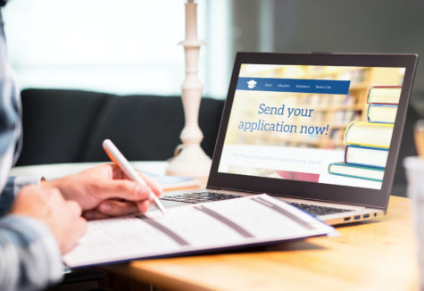 A man filling out a paper application, while sitting in front of a laptop that states "Send your application now!".