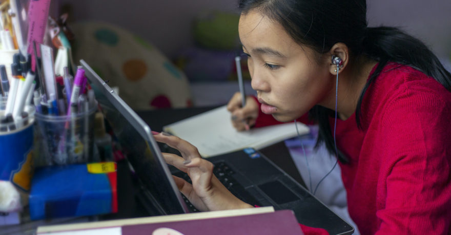 A female student of Asian descent studying hard at her laptop while taking notes in a notebook.