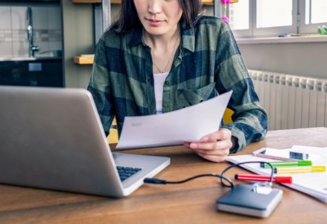 A woman working in front of a laptop, surrounded by papers, colored pens, and an external hard-drive.