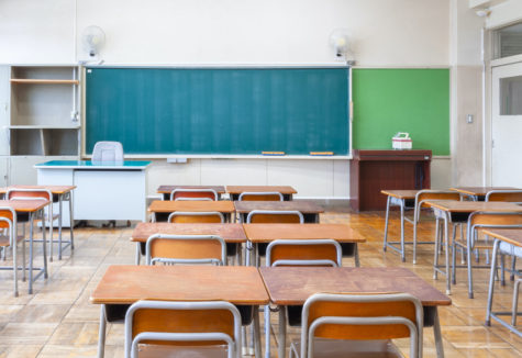 The inside of an empty classroom with a chalkboard and wooden-topped desks.