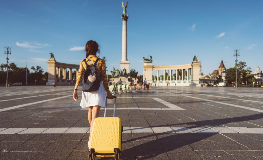 A female pulling her yellow rolling suitcase across a town square in a foreign city.