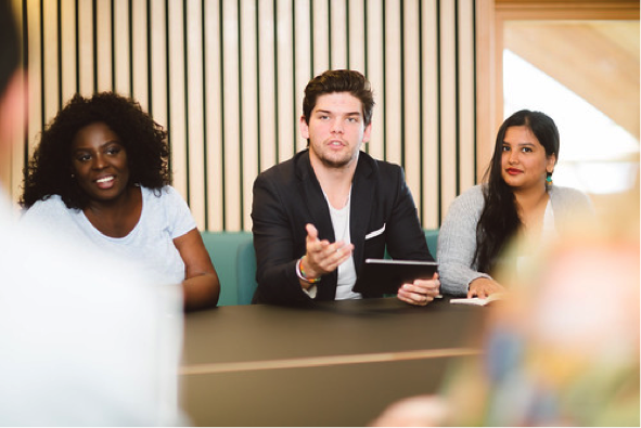 Three students, two female one male, presenting something to a group from across a conference table.