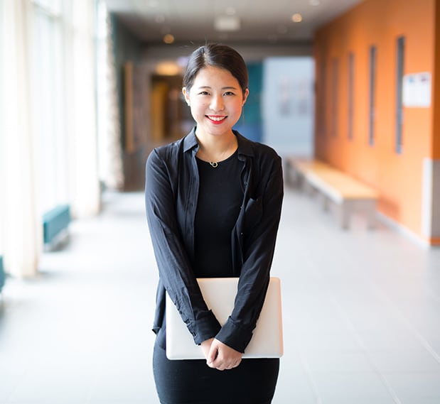 Smiling female teen of Asian descent standing in school hallway holding laptop near waist