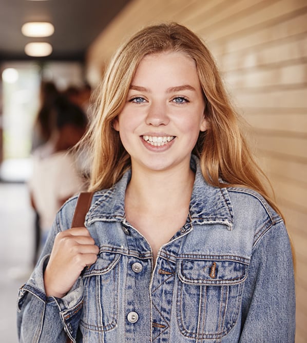 Smiling young blonde high school girl standing in hallway at school with other students behind her