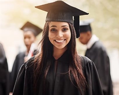 Smiling high school girl of Indian descent wearing graduation cap and gown with classmates in background