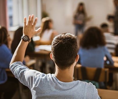 High school boy seated at classroom desk raising hand